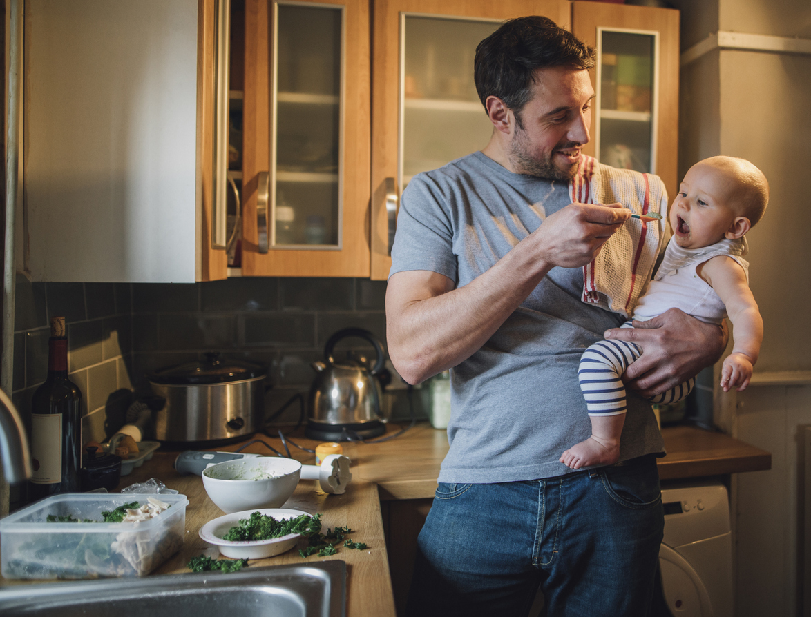 Father is standing in the kitchen of his home with his baby in his arms. He is feeding him with a spoon and spinach and vegetables can be seen on the worktop with a blender.
